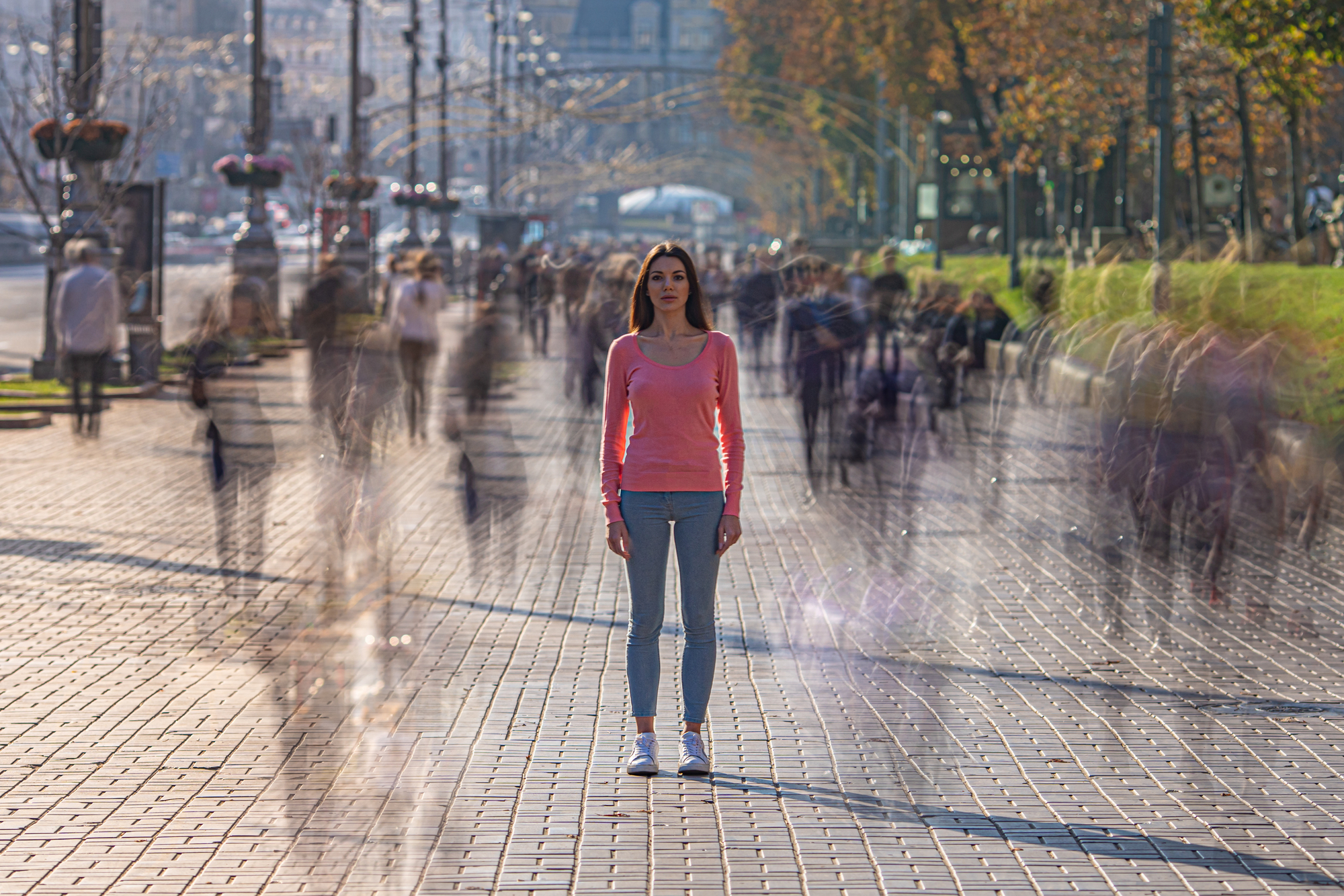 A woman in a pink shirt and blue jeans stands still in the middle of a busy pedestrian walkway. The people around her are blurred, conveying a sense of motion and speed as they walk past. The background includes trees and cityscape elements.