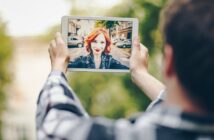 A person takes a selfie using a tablet outdoors. The tablet screen shows a woman with red hair, wearing a black leather jacket and bright red lipstick, smiling at the camera. The background features a city street with parked cars and trees.