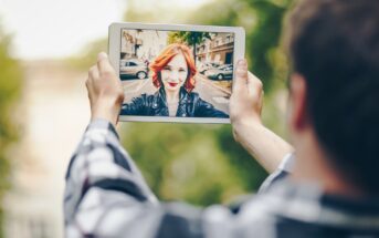 A person takes a selfie using a tablet outdoors. The tablet screen shows a woman with red hair, wearing a black leather jacket and bright red lipstick, smiling at the camera. The background features a city street with parked cars and trees.