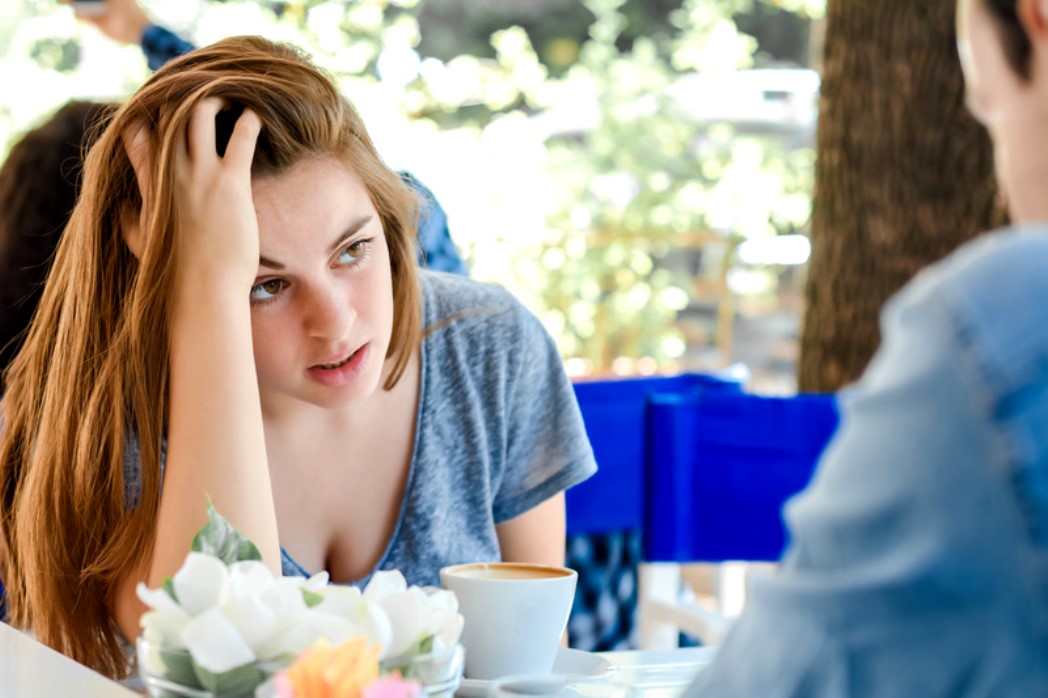 A woman with brown hair rests her head on her hand, looking intently at another person, who is not fully visible, during a conversation at an outdoor café. A coffee cup and a small vase of flowers are on the table in front of her.