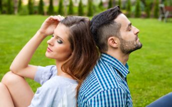 A woman and a man are sitting back-to-back on a grassy lawn. The woman, with long brown hair, has her eyes closed and is leaning her head slightly to one side with her hand resting on her head. The man, with short dark hair and a beard, also has his eyes closed.