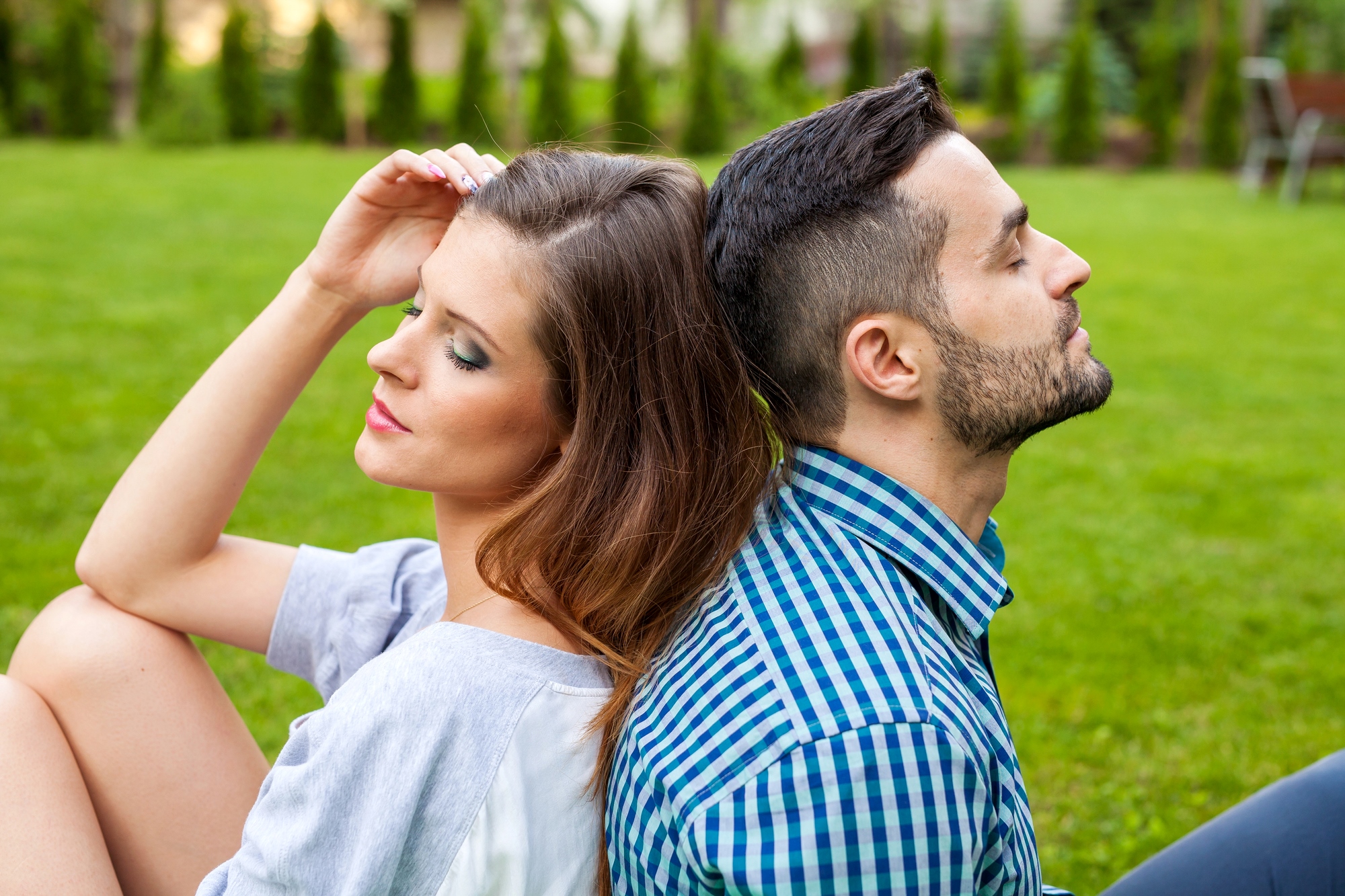 A woman and a man are sitting back-to-back on a grassy lawn. The woman, with long brown hair, has her eyes closed and is leaning her head slightly to one side with her hand resting on her head. The man, with short dark hair and a beard, also has his eyes closed.