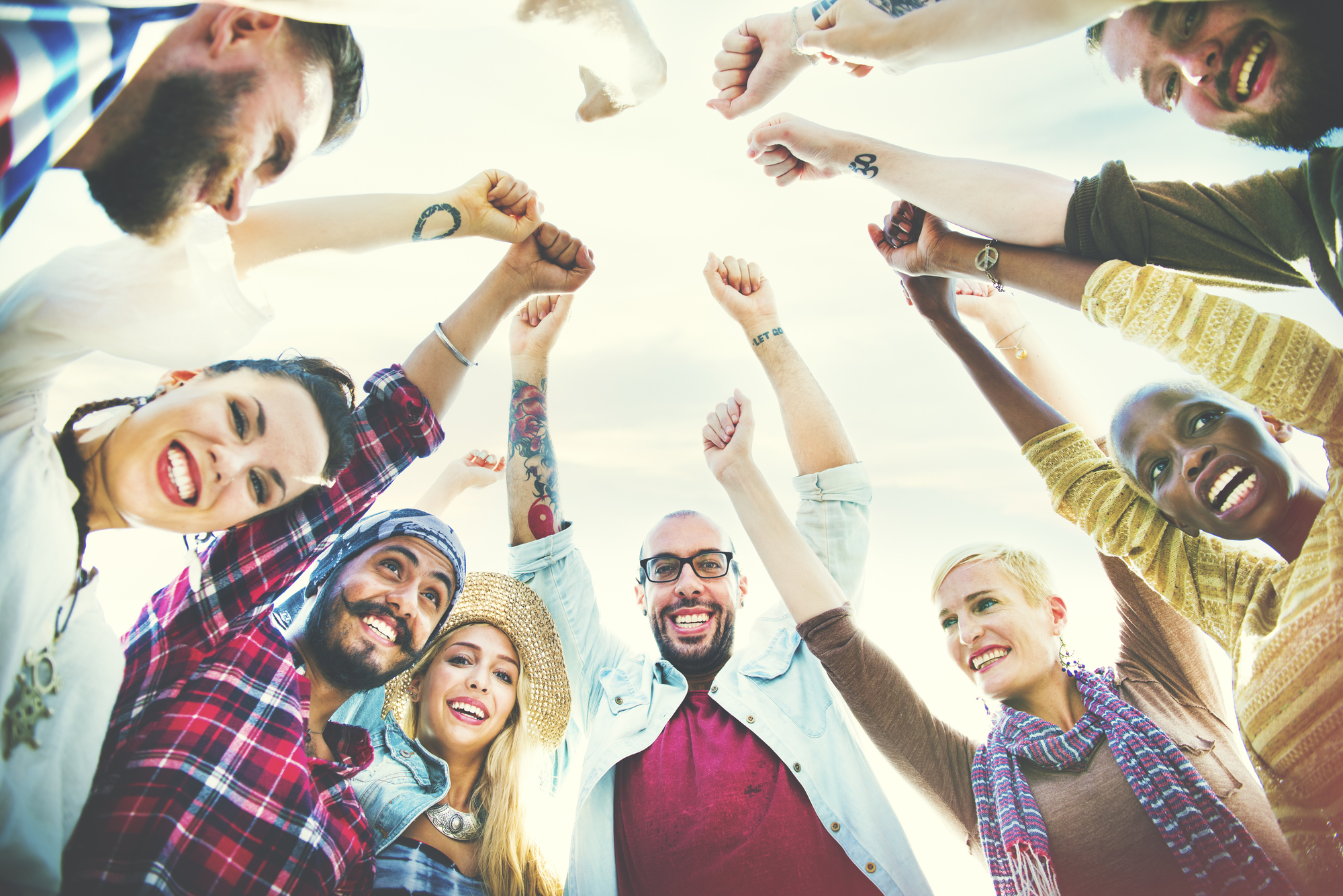 A diverse group of people stands in a circle with their hands raised and fists clenched, smiling joyfully. They appear cheerful and united, captured from a low-angle view against a clear sky backdrop. They are casually dressed, showcasing various styles.