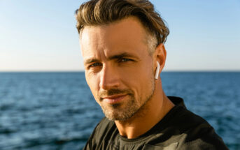 A man with short, styled hair and a trimmed beard is standing by the sea, wearing a black shirt and white earbuds. He is looking directly at the camera, and the ocean and sky are in the background.