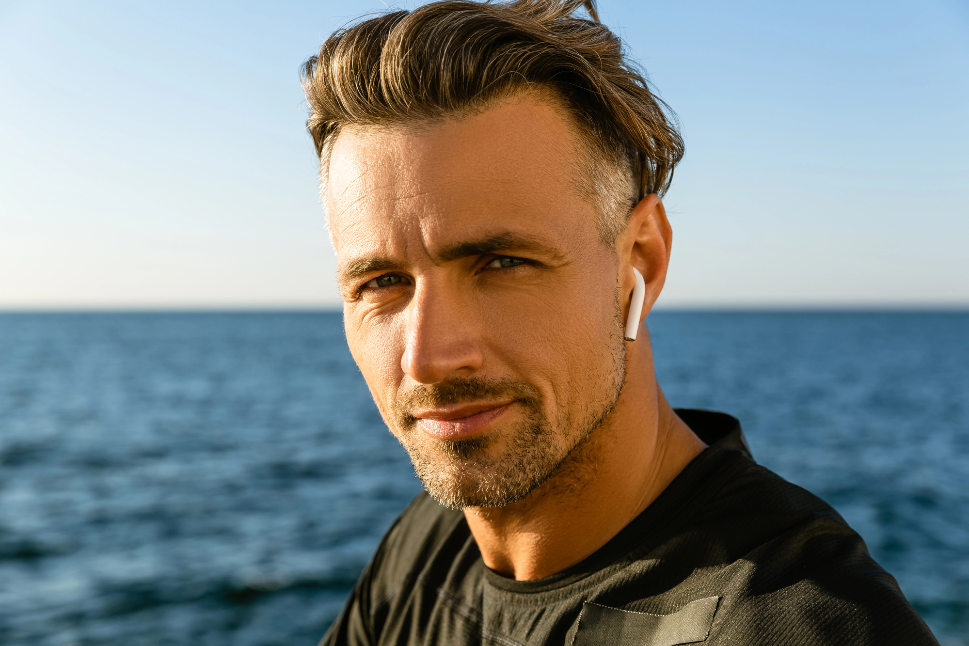 A man with short, styled hair and a trimmed beard is standing by the sea, wearing a black shirt and white earbuds. He is looking directly at the camera, and the ocean and sky are in the background.