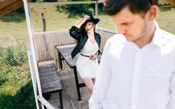 A woman in a white dress and blue hat poses confidently under a wooden gazebo, her hand on her hat. In the foreground, a man in a white shirt looks down, seemingly distant. The gazebo is set in a grassy, fenced outdoor area.