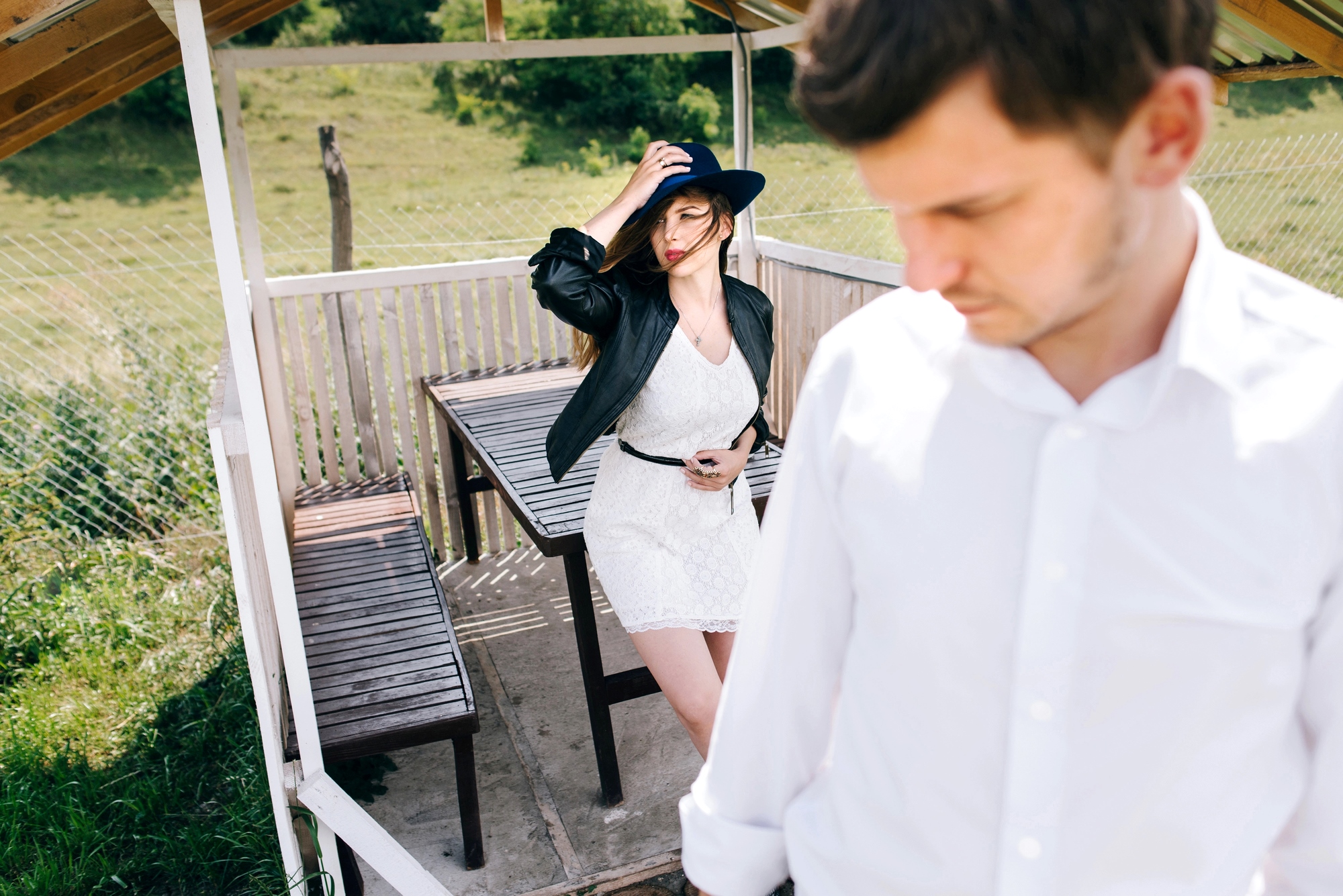 A woman in a white dress and blue hat poses confidently under a wooden gazebo, her hand on her hat. In the foreground, a man in a white shirt looks down, seemingly distant. The gazebo is set in a grassy, fenced outdoor area.