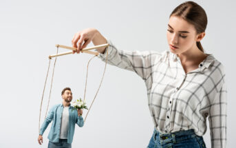 A woman in a plaid shirt holds puppet strings attached to a smaller man holding white flowers, making him appear as a puppet. The man is smiling and looking up at the woman. Both are set against a plain, light background.