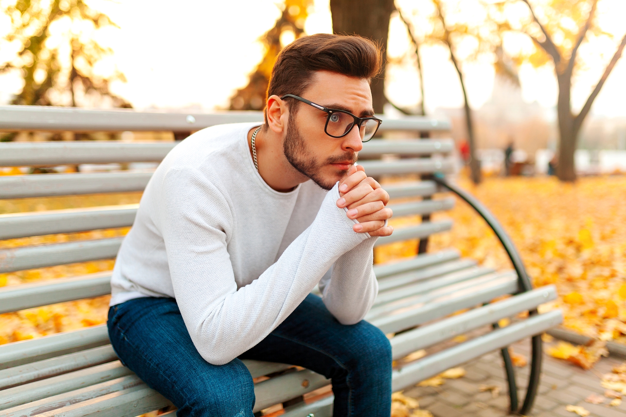 A man with glasses, wearing a white sweater and blue jeans, sits on a park bench in an autumn setting. He is resting his chin on his clasped hands, looking thoughtful. Fallen leaves surround the bench and trees with yellow foliage are visible in the background.