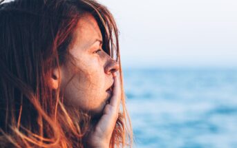 A woman with long, auburn hair looks pensively into the distance with her hand resting on her chin. Behind her, a blurred seascape under a clear sky is visible, capturing a serene, contemplative moment by the ocean.