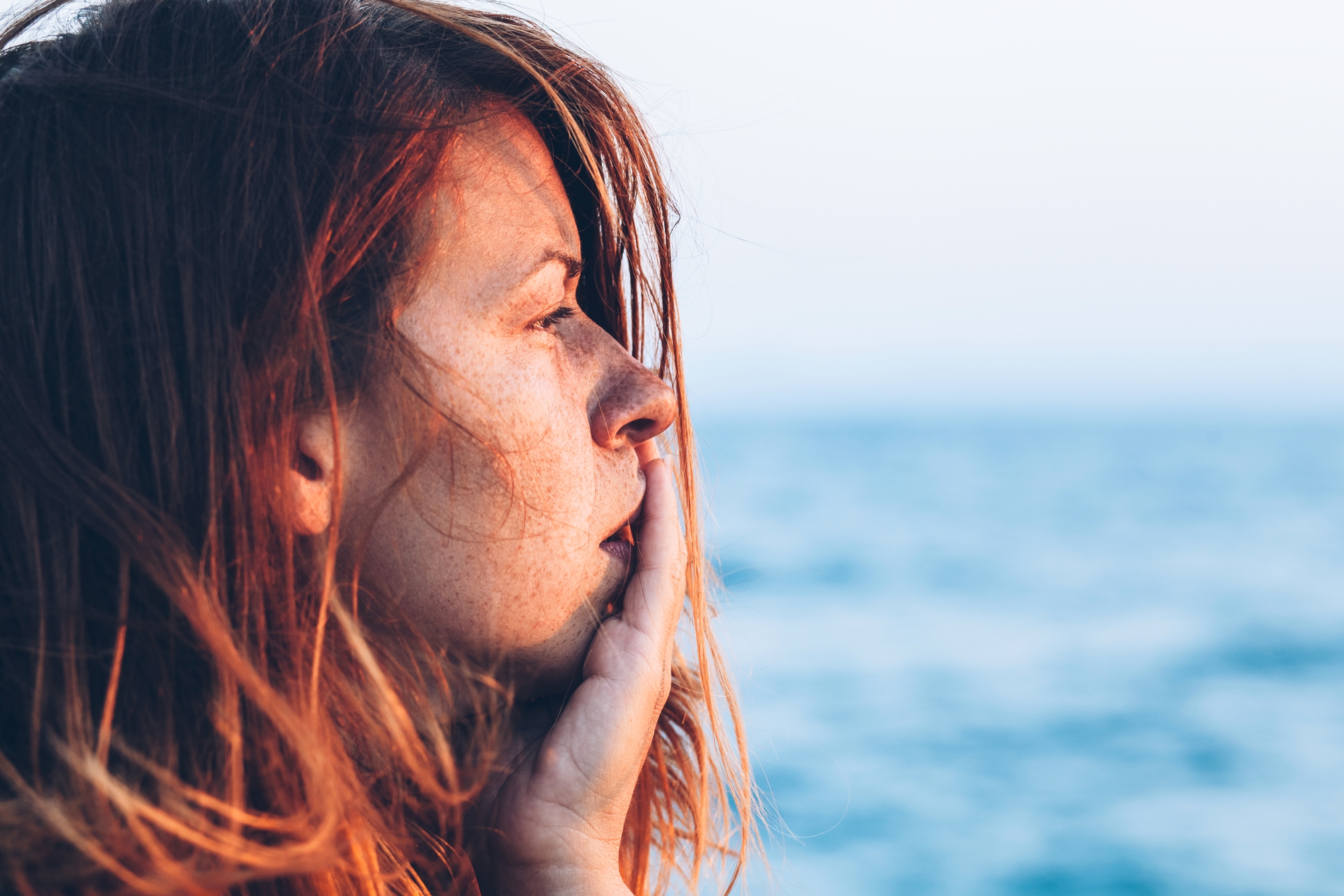 A woman with long, auburn hair looks pensively into the distance with her hand resting on her chin. Behind her, a blurred seascape under a clear sky is visible, capturing a serene, contemplative moment by the ocean.