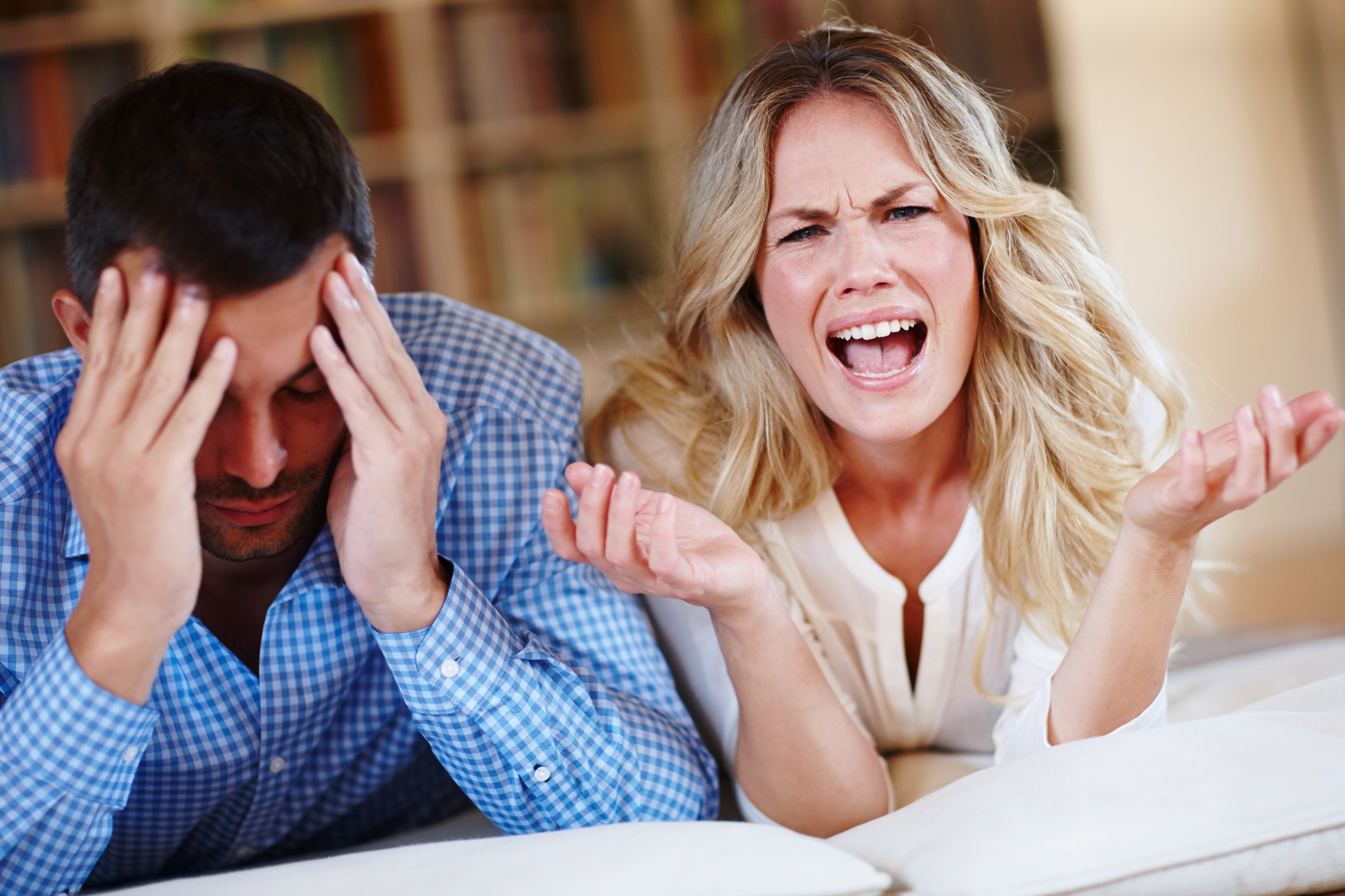 A woman with long blonde hair is sitting next to a man with short dark hair. She appears frustrated or upset, gesturing with one hand. The man has his head in his hands, looking down. They are indoors, with bookshelves blurred in the background.