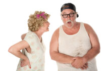 An elderly woman with curly hair and a pink bow, wearing a floral dress, appears to be scolding a bearded man in a dirty tank top, glasses, and a backwards cap. The man looks surprised and holds his hands near his stomach. They stand against a white background.