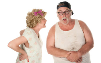 An elderly woman with curly hair and a pink bow, wearing a floral dress, appears to be scolding a bearded man in a dirty tank top, glasses, and a backwards cap. The man looks surprised and holds his hands near his stomach. They stand against a white background.