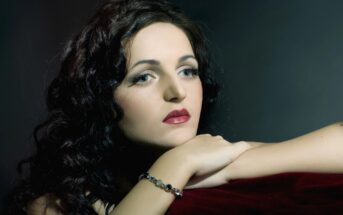 A person with long, wavy dark hair and red lipstick, wearing a gemstone bracelet, is resting their arms and head on a red velvet surface. The background is dark, creating a dramatic contrast with the person's light skin and contemplative expression.
