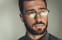 A close-up of a man with brown hair and a beard wearing round glasses and a serious expression. He has a contemplative look in his eyes and is dressed in a dark shirt, with a blurred neutral background.