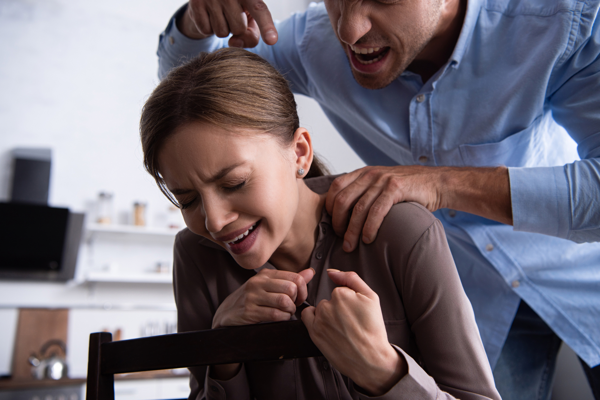 A woman sits in a chair, looking distressed and upset, while a man stands behind her, gripping her shoulders and shouting aggressively. Both are indoors in a kitchen setting, with blurry shelves and items visible in the background.