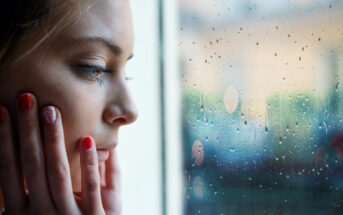 Close-up of a person with long hair looking out a window with raindrops on it. They are touching their face with both hands, and their expression appears contemplative. The background is blurred. Their nails are painted red, with a decorative design on one nail.