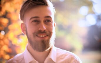 A man with light brown hair, a beard, and wearing a white shirt is smiling warmly. The background is softly blurred with autumnal colors, suggesting a sunny, outdoor setting. The light creates a gentle, glowing effect around the man.