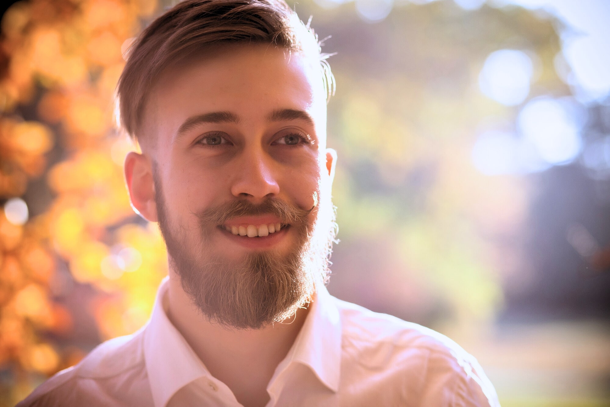 A man with light brown hair, a beard, and wearing a white shirt is smiling warmly. The background is softly blurred with autumnal colors, suggesting a sunny, outdoor setting. The light creates a gentle, glowing effect around the man.