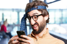 A man with glasses and a beard is wearing a blue tie wrapped around his head like a headband. He is looking at his smartphone with a humorous, exaggeratedly confused expression. The background is a blurry indoor space with large windows.