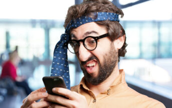 A man with glasses and a beard is wearing a blue tie wrapped around his head like a headband. He is looking at his smartphone with a humorous, exaggeratedly confused expression. The background is a blurry indoor space with large windows.