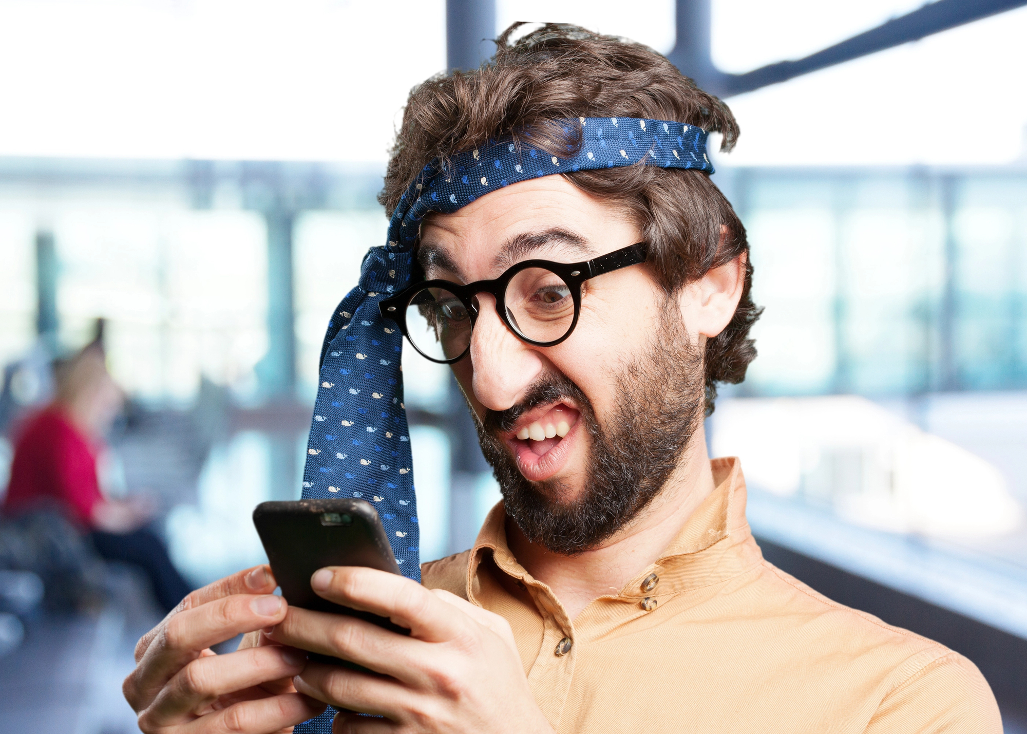 A man with glasses and a beard is wearing a blue tie wrapped around his head like a headband. He is looking at his smartphone with a humorous, exaggeratedly confused expression. The background is a blurry indoor space with large windows.