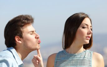 A young man with his eyes closed puckers his lips, attempting to kiss a young woman. The woman, with long brown hair and wearing a blue sleeveless top, turns her head away, avoiding the kiss. The background is blurred, with a clear sky and distant landscape.