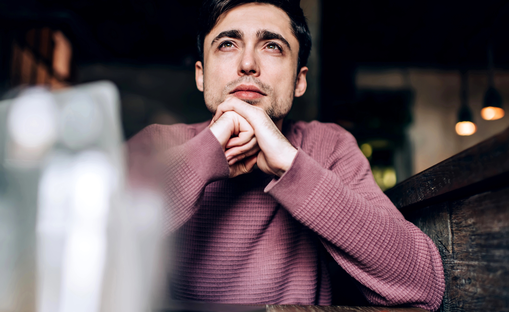 A man with short dark hair is sitting at a wooden table indoors, resting his chin on his clasped hands and looking thoughtfully into the distance. He is wearing a pink knit sweater, and the background is softly blurred with warm lighting.