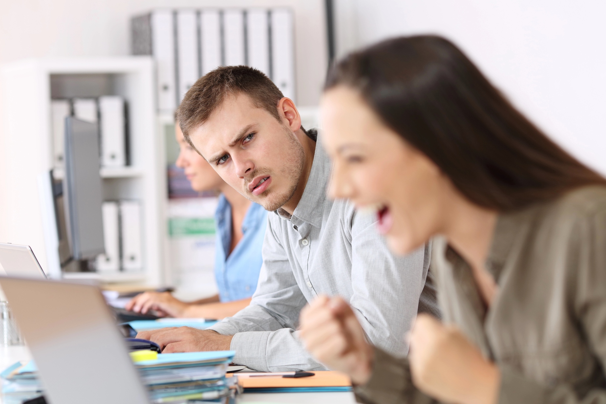 In an office setting, a woman in the foreground cheers enthusiastically at her laptop, while a man next to her looks puzzled and concerned. Another person is visible in the background, working on a computer. Shelves with binders are seen in the background.