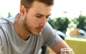 A man with short brown hair and a beard, wearing a grey t-shirt, is sitting at a table outdoors. He appears to be deep in thought, looking down at something out of the frame. The background is blurred with greenery and a patio setting.