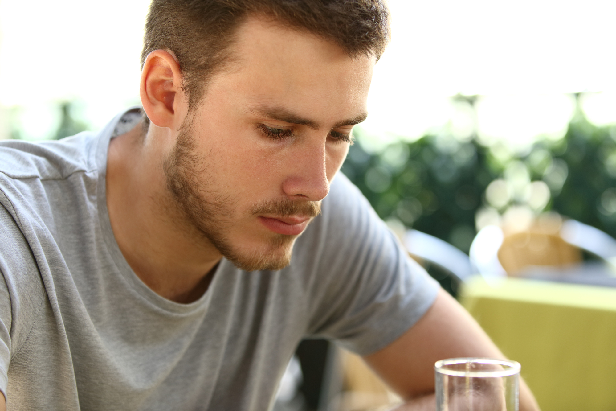 A man with short brown hair and a beard, wearing a grey t-shirt, is sitting at a table outdoors. He appears to be deep in thought, looking down at something out of the frame. The background is blurred with greenery and a patio setting.