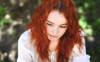 A person with long, curly red hair is looking down thoughtfully. They are wearing a white shirt and are outside with a blurred, natural green background. The sunlight illuminates their hair and face gently.