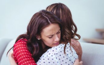 Two women are embraced in a comforting hug. The woman facing the camera has long brown hair and is wearing a red patterned blouse. Her eyes are closed with a pained expression. The other woman, with brown hair and a white blouse with black patterns, is turned away.