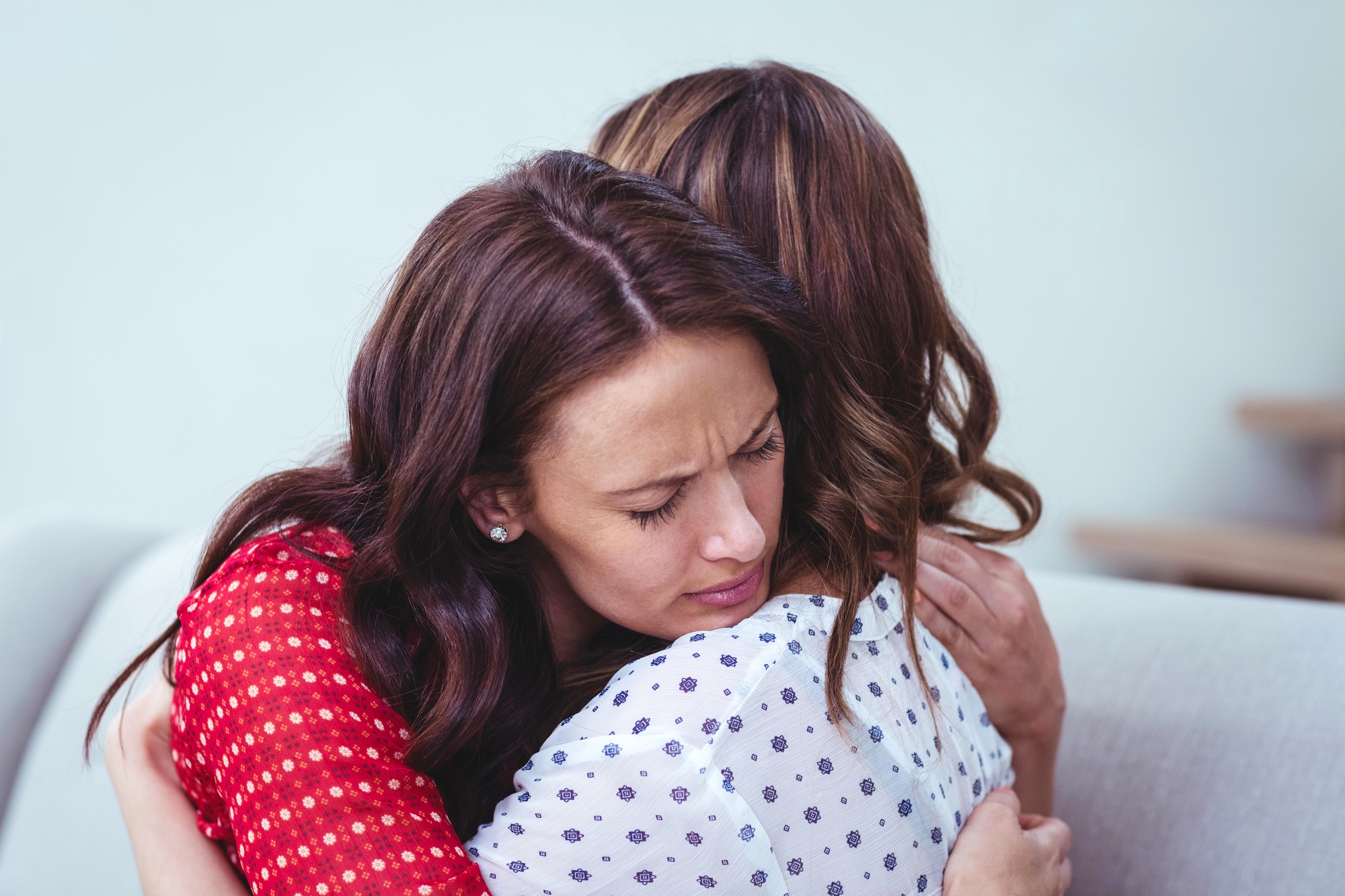 Two women are embraced in a comforting hug. The woman facing the camera has long brown hair and is wearing a red patterned blouse. Her eyes are closed with a pained expression. The other woman, with brown hair and a white blouse with black patterns, is turned away.