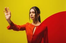 A woman with long dark hair stands against a vibrant yellow background with a red curved shape. She is wearing a red top and has one hand raised in front of her, creating a dynamic and striking visual composition.