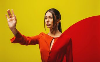 A woman with long dark hair stands against a vibrant yellow background with a red curved shape. She is wearing a red top and has one hand raised in front of her, creating a dynamic and striking visual composition.