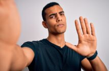 A man with short black hair and a black t-shirt is taking a selfie. He is raising his right hand, palm facing the camera, as if gesturing to stop or hold. His expression appears serious. The background is plain and neutral.