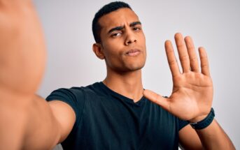 A man with short black hair and a black t-shirt is taking a selfie. He is raising his right hand, palm facing the camera, as if gesturing to stop or hold. His expression appears serious. The background is plain and neutral.