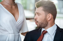 A man in a suit and red tie appears to be making a surprised or impressed facial expression, looking at the chest of a woman in a white dress with a low neckline. The background is blurred with natural light coming through windows.