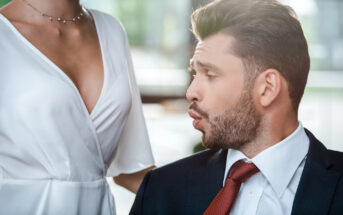 A man in a suit and red tie appears to be making a surprised or impressed facial expression, looking at the chest of a woman in a white dress with a low neckline. The background is blurred with natural light coming through windows.