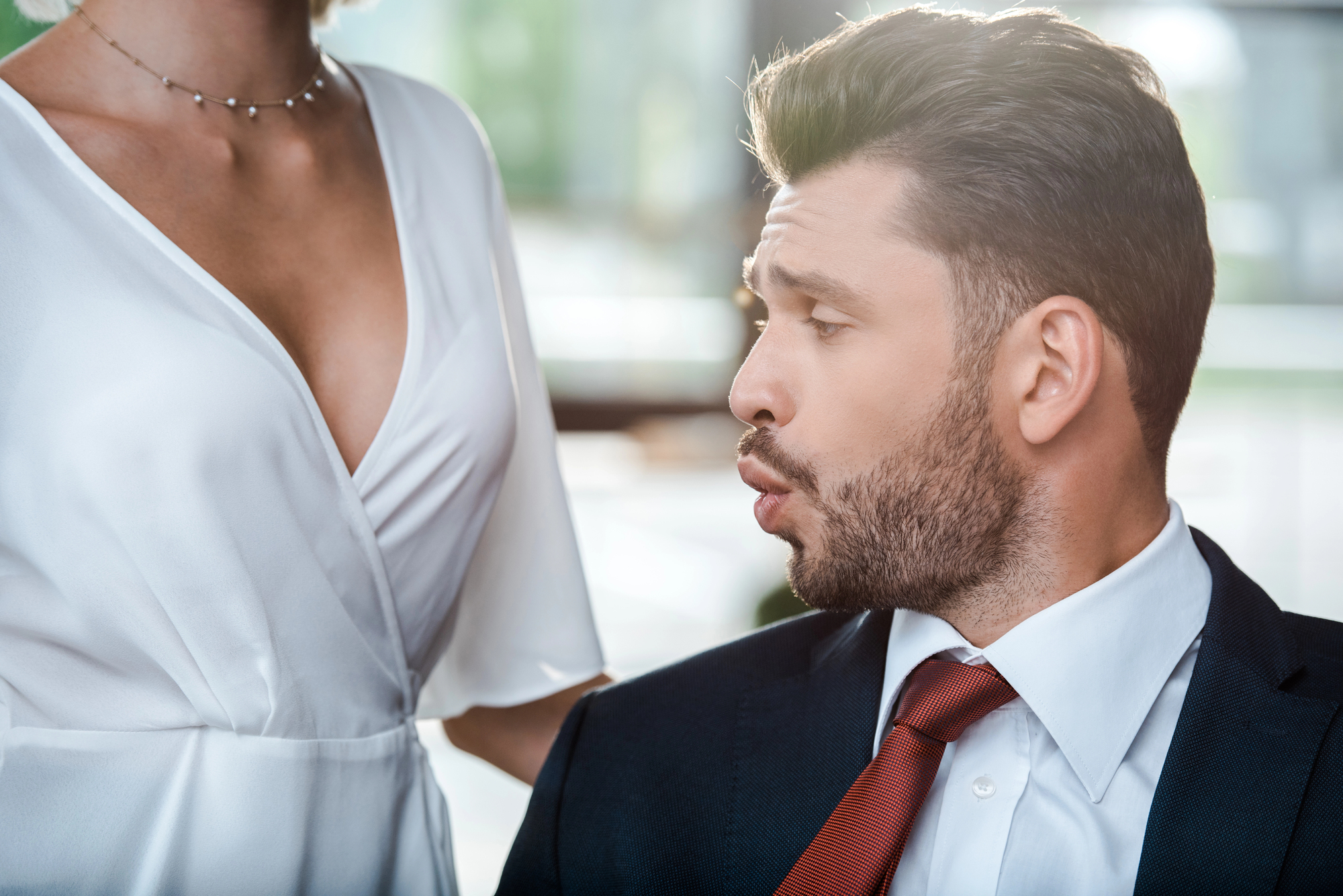 A man in a suit and red tie appears to be making a surprised or impressed facial expression, looking at the chest of a woman in a white dress with a low neckline. The background is blurred with natural light coming through windows.