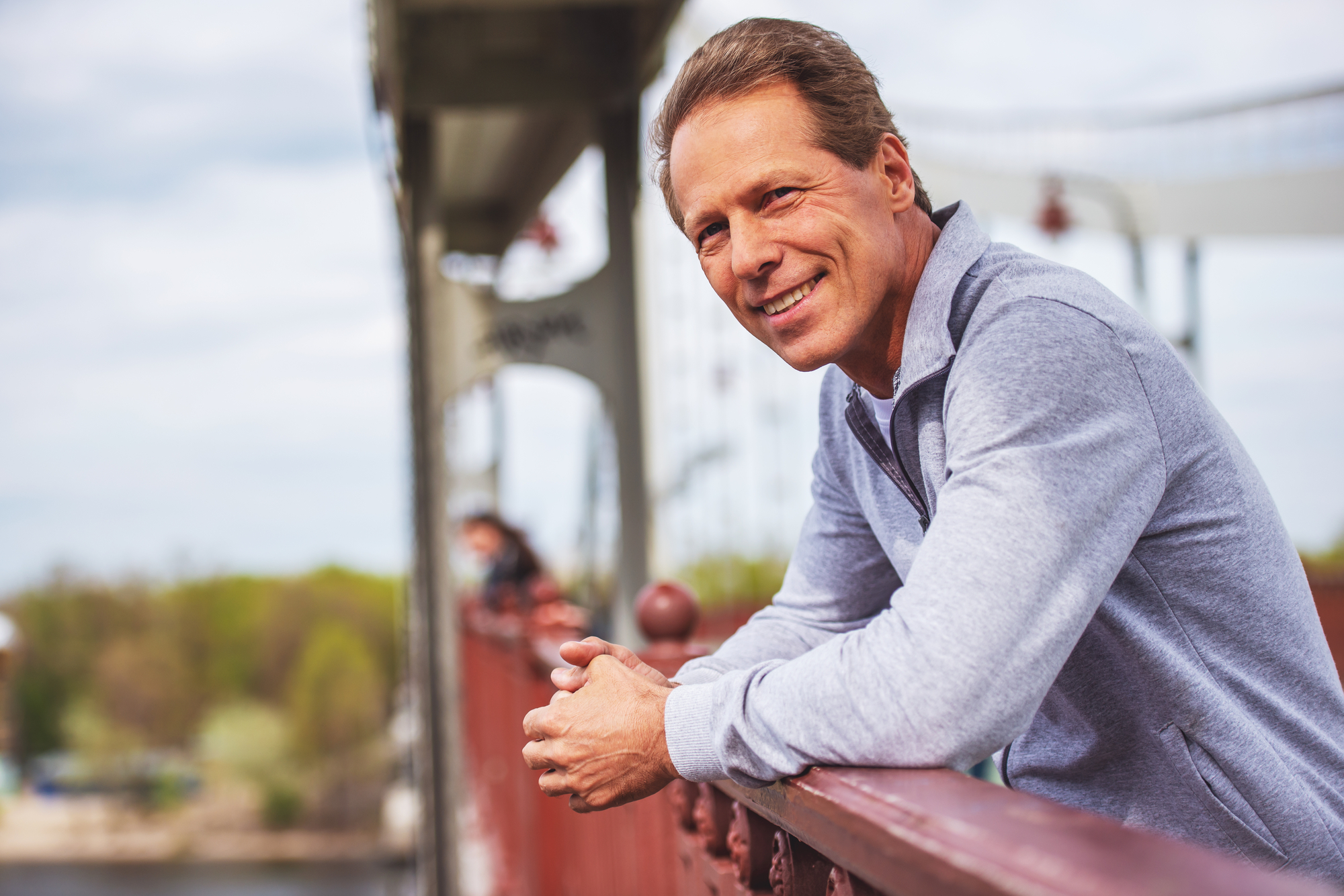 A smiling man in a light gray sweater leans on the railing of a bridge. The bridge structure is visible behind him, and there is a scenic view with greenery and water in the background. The sky is slightly overcast.