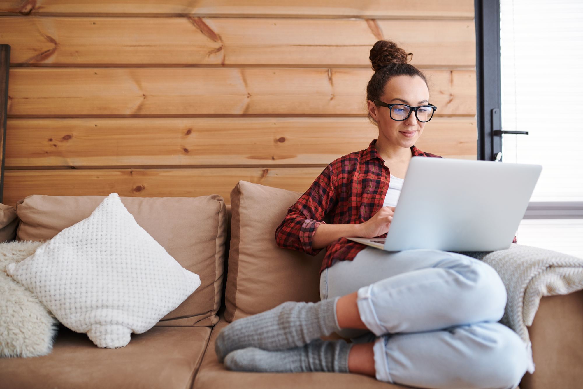 A person with a bun hairstyle and glasses is sitting on a beige couch with fluffy cushions, wearing a red plaid shirt and light blue pants. They are working on an open laptop in a cozy room with wooden paneled walls and soft natural light.