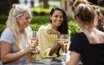 Four women are sitting at an outdoor table, smiling and talking while holding glasses of rosé wine. The setting appears to be a sunny day in a park or garden, with greenery in the background.