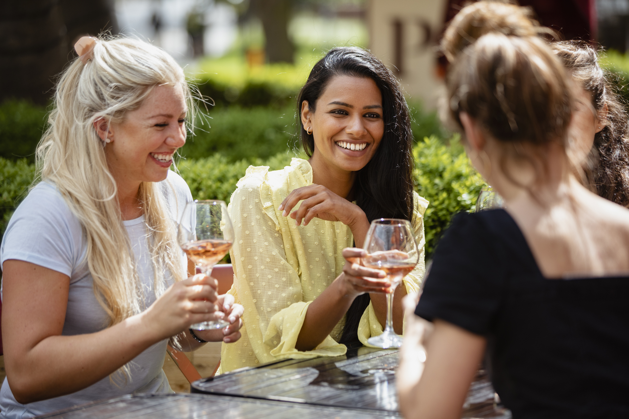 Four women are sitting at an outdoor table, smiling and talking while holding glasses of rosé wine. The setting appears to be a sunny day in a park or garden, with greenery in the background.