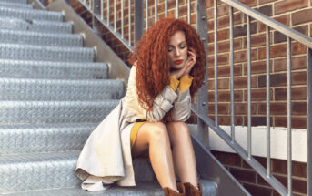 A woman with long, curly red hair sits on an outdoor metal staircase, leaning on the railing. She wears a light-colored coat over a yellow dress, and brown boots. Her head rests on her hands, and she appears thoughtful. A brick wall is in the background.