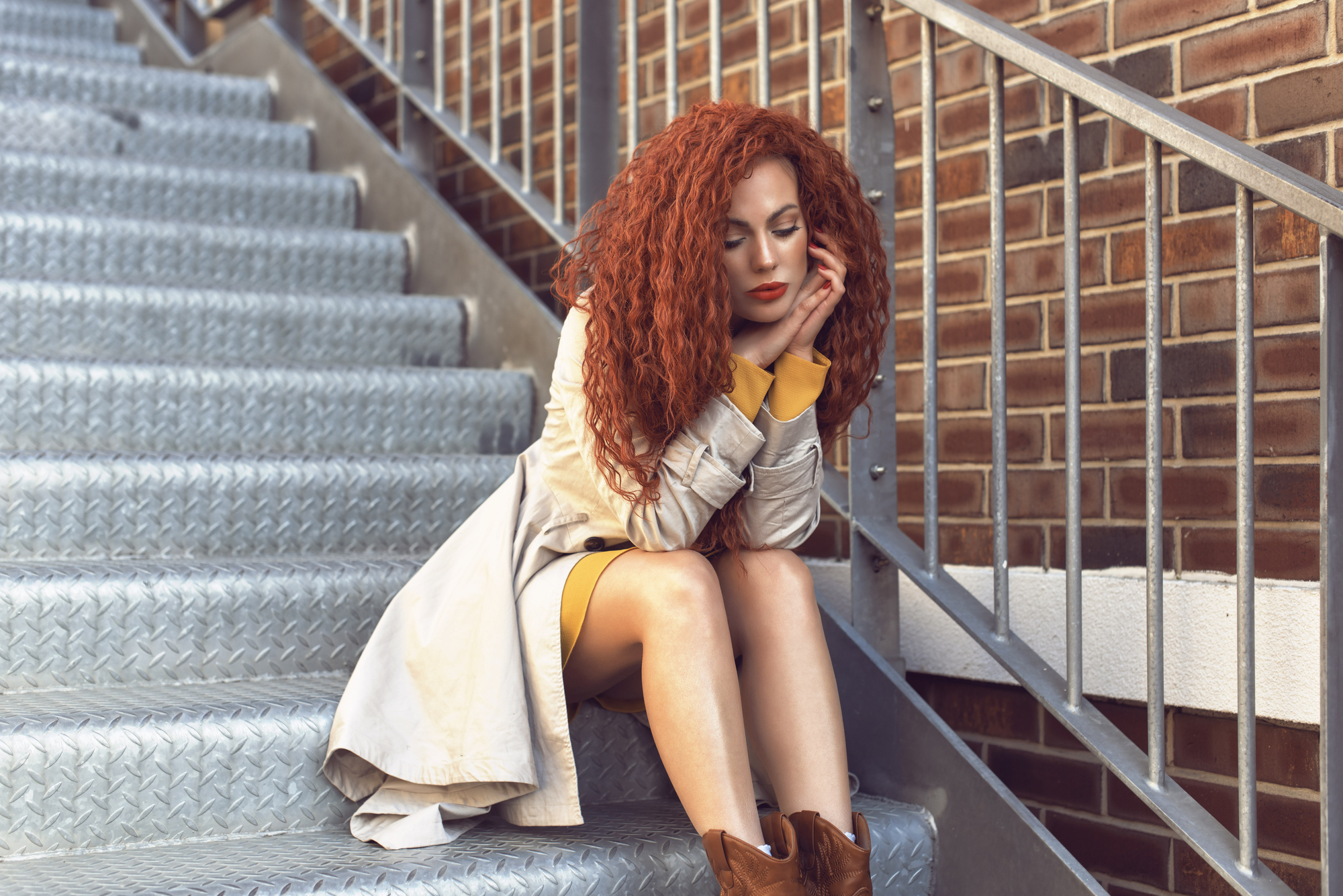 A woman with long, curly red hair sits on an outdoor metal staircase, leaning on the railing. She wears a light-colored coat over a yellow dress, and brown boots. Her head rests on her hands, and she appears thoughtful. A brick wall is in the background.