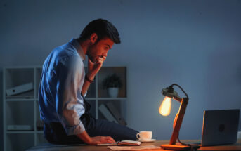 A man in a shirt and tie sits on a desk, looking pensive with his hand on his chin. A lit desk lamp, cup of coffee, and open notebook are in front of him. He is working late in a dimly lit office. A laptop is to the side, and shelves of binders are behind him.