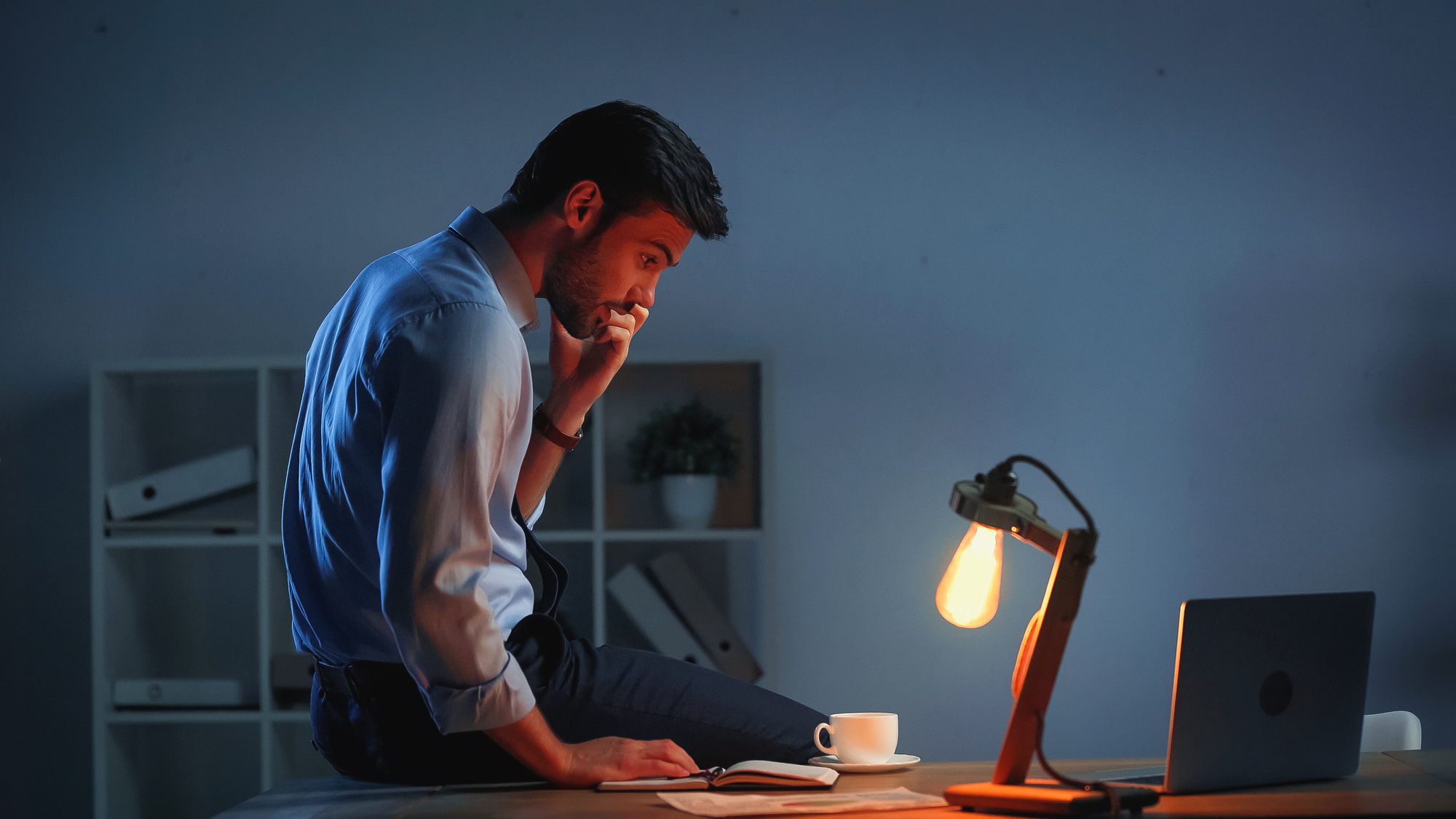 A man in a shirt and tie sits on a desk, looking pensive with his hand on his chin. A lit desk lamp, cup of coffee, and open notebook are in front of him. He is working late in a dimly lit office. A laptop is to the side, and shelves of binders are behind him.